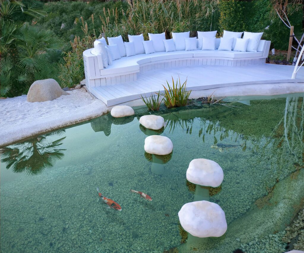 Piscina natural elegante con paso blanco y peces koi en nuestro diseño de jardín mediterráneo.